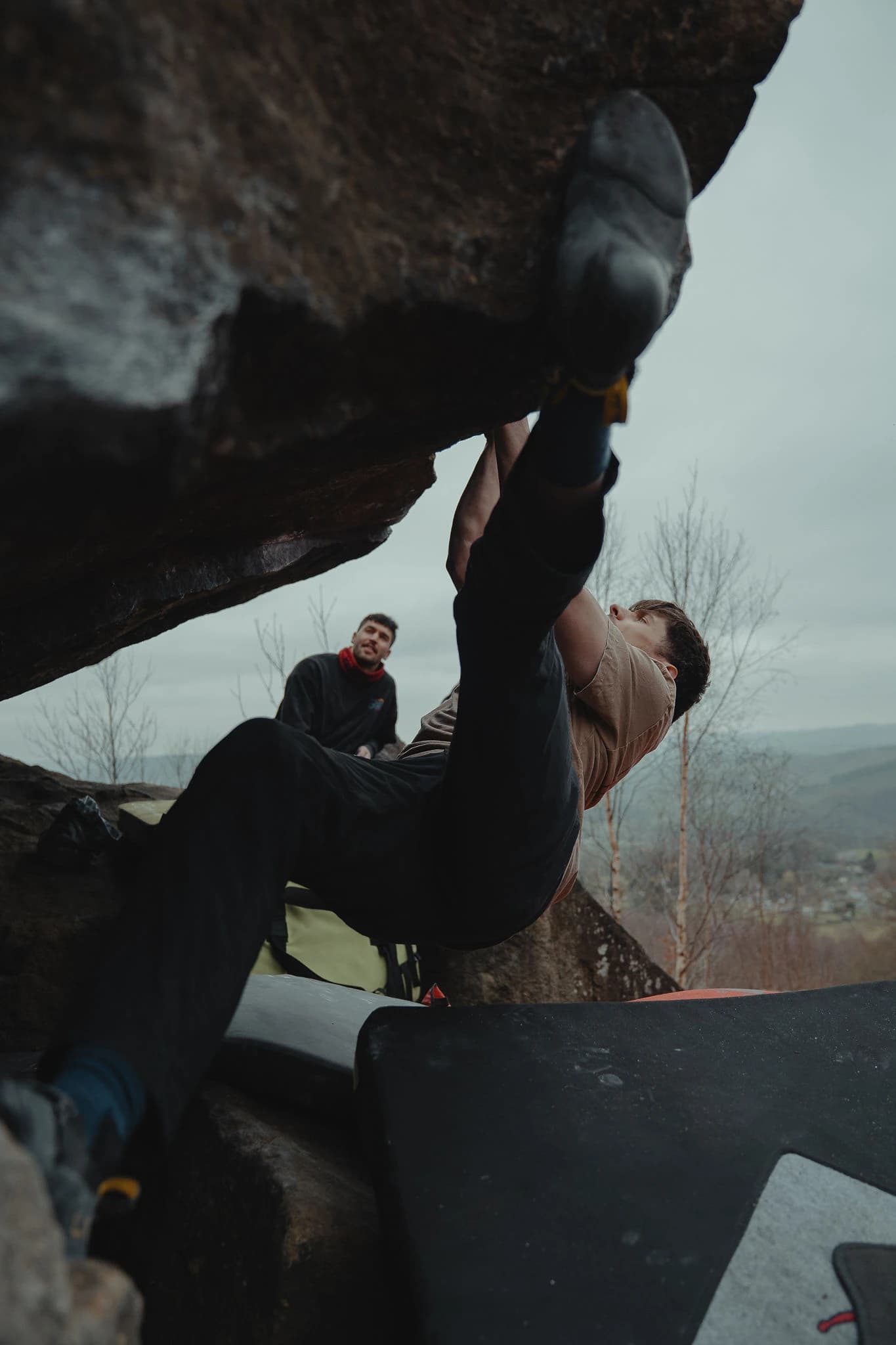 Climber working an outdoor overhang with a spotter and crash pads below